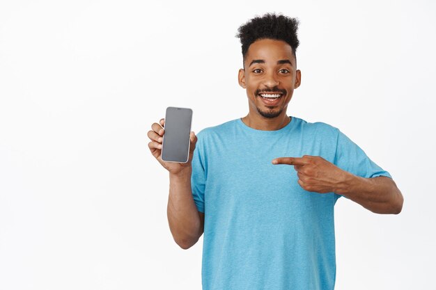 Porträt eines gutaussehenden, glücklichen afroamerikanischen Mannes, der mit dem Finger auf das Handy zeigt, erfreut lächelt, Smartphone-Werbung zeigt, Laden oder App empfiehlt, weißer Hintergrund
