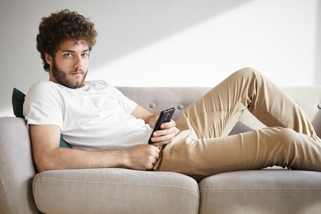 Porträt eines gutaussehenden jungen europäischen Mannes mit haarigem Gesicht, das sich auf bequemem Sofa ausruht, Newsfeed über soziale Netzwerke auf seinem Handy durchsucht, Beiträge mag und Kommentare online hinterlässt