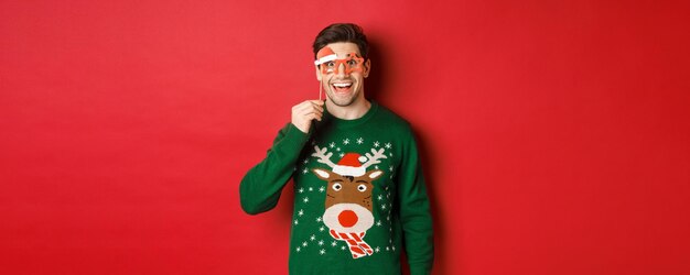 Porträt eines gutaussehenden lächelnden Mannes in Weihnachtspullover und Partybrille, der überrascht und glücklich aussieht