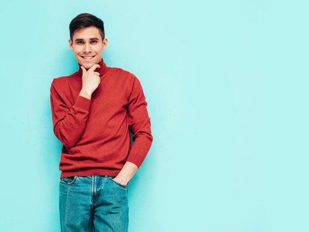 Porträt eines gutaussehenden, lächelnden Modells Sexy stilvoller Mann in rotem Rollkragenpullover und Jeans Mode-Hipster-Männchen posiert in der Nähe der blauen Wand im Studio Isoliert