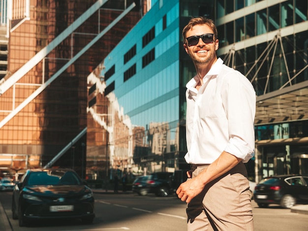 Porträt eines gutaussehenden, lächelnden, stilvollen Hipster-Lambersexual-Modells. Moderner Mann in weißem Hemd. Modemann, der im Straßenhintergrund in der Nähe von Wolkenkratzern mit Sonnenbrille posiert