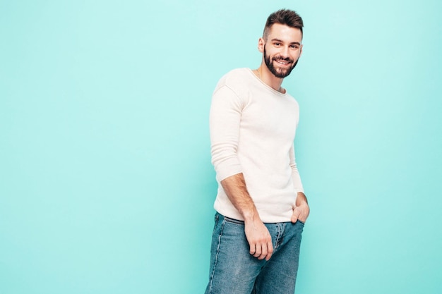 Porträt eines gutaussehenden, lächelnden, stilvollen Hipster-Lambersexual-ModelsMann in weißem Pullover und Jeans Modemann isoliert auf blauer Wand im Studio