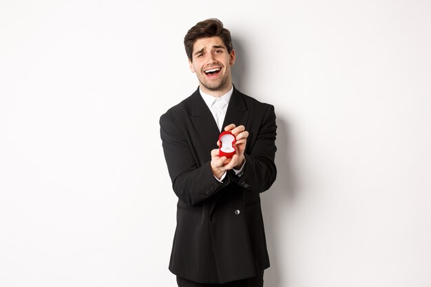 Porträt eines gutaussehenden Mannes im schwarzen Anzug, offene Kiste mit Ehering, der einen Vorschlag macht, ihn heiraten möchte und vor weißem Hintergrund steht.