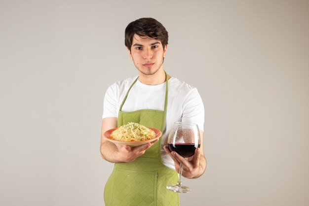 Porträt eines gutaussehenden Mannes in Schürze, der einen Teller mit Nudeln und ein Glas Wein hält.