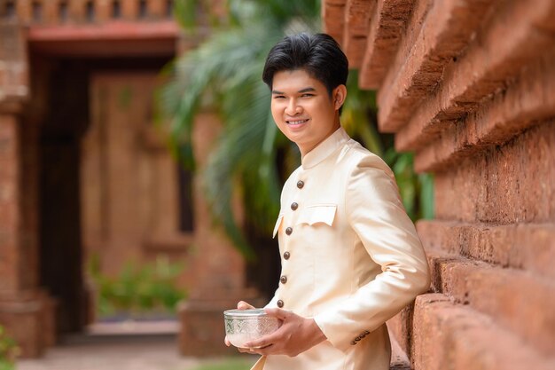 Porträt eines gutaussehenden Mannes in wunderschönen thailändischen Kostümen, der mit einer Wasserschüssel im Tempel am Songkran-Festival am thailändischen Neujahrsfamilientag im April steht