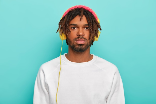 Porträt eines gutaussehenden selbstbewussten Mannes mit Dreadlocks, trägt Kopfhörer, die an ein Gerät angeschlossen sind, hört warme Musik oder Radio, genießt eine gemütliche Atmosphäre, trägt einen weißen Freizeitpullover und rosige Kopfbedeckungen
