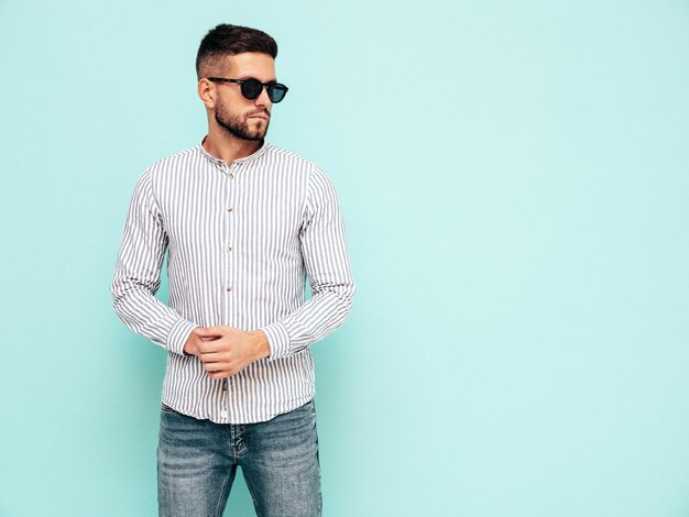 Porträt eines gutaussehenden, selbstbewussten Modells Sexy stilvoller Mann in Hemd und Jeans Mode-Hipster-Mann posiert in der Nähe der blauen Wand im Studio mit Sonnenbrille