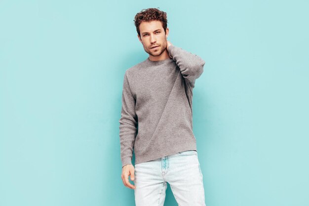 Porträt eines gutaussehenden, selbstbewussten Modells Sexy stilvoller Mann in Pullover und Jeans Mode-Hipster-Mann mit lockiger Frisur posiert in der Nähe der blauen Wand im Studio Isoliert