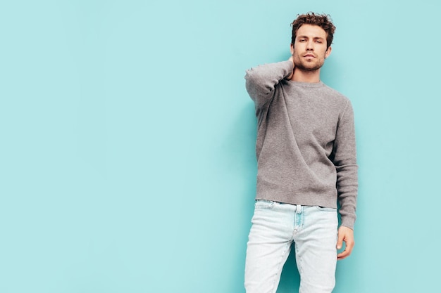 Porträt eines gutaussehenden, selbstbewussten Modells Sexy stilvoller Mann in Pullover und Jeans Mode-Hipster-Mann mit lockiger Frisur posiert in der Nähe der blauen Wand im Studio Isoliert