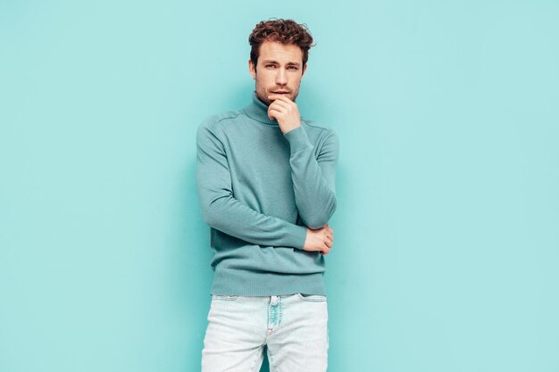 Porträt eines gutaussehenden, selbstbewussten Modells Sexy stilvoller Mann in Pullover und Jeans Mode-Hipster-Mann mit lockiger Frisur posiert in der Nähe der blauen Wand im Studio Isoliert