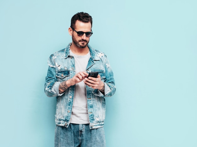 Porträt eines gutaussehenden, selbstbewussten, stilvollen Hipster-Lambersexual-Modells Sexy Mann in Jeansjacke gekleidet Mode männlich isoliert auf blauer Wand im Studio mit Sonnenbrille