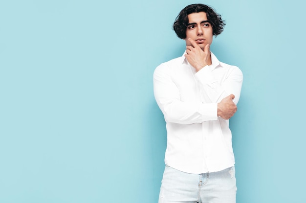 Porträt eines gutaussehenden, selbstbewussten, stilvollen Hipster-Lambersexual-Modells Sexy Mann in sommerlichem weißem Hemd und Jeanskleidung Mode männlich isoliert im Studio Posiert in der Nähe der blauen Wand