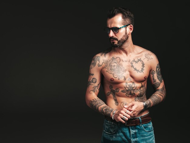 Porträt eines gutaussehenden, selbstbewussten, stilvollen Hipster-Lambersexual-Modells. Sexy moderner Mann. Nackter Oberkörper mit Tätowierungen. Modemann, der im Studio auf dunklem Hintergrund mit Sonnenbrille posiert