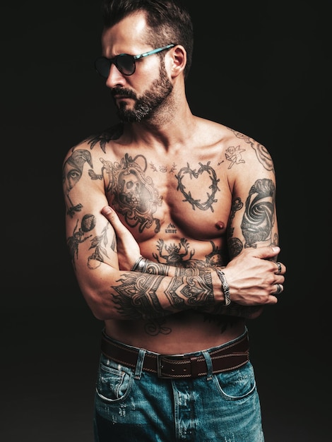 Porträt eines gutaussehenden, selbstbewussten, stilvollen Hipster-Lambersexual-Modells. Sexy moderner Mann. Nackter Oberkörper mit Tätowierungen. Modemann, der im Studio auf dunklem Hintergrund mit Sonnenbrille posiert