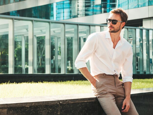 Porträt eines gutaussehenden, selbstbewussten, stilvollen Hipster-Lambersexual-ModellsModerner Mann in weißem Hemd Modemann, der im Straßenhintergrund in der Nähe von Wolkenkratzern mit Sonnenbrille im Freien bei Sonnenuntergang posiert