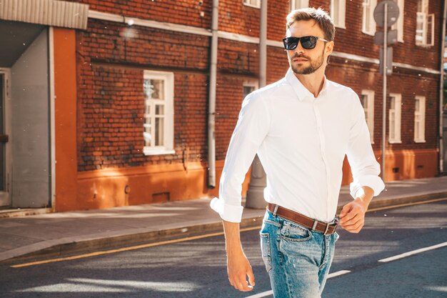 Porträt eines gutaussehenden, selbstbewussten, stilvollen Hipster-Lambersexual-ModellsModerner Mann in weißem Hemd Modemann posiert auf dem Straßenhintergrund mit Sonnenbrille Im Freien bei Sonnenuntergang