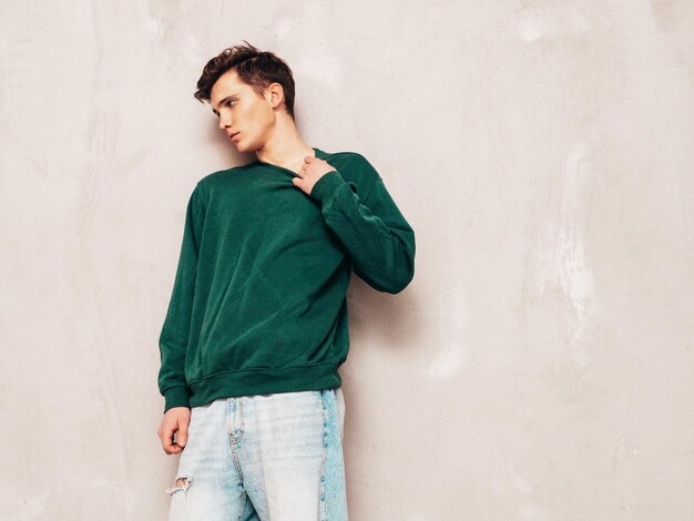 Porträt eines gutaussehenden, selbstbewussten, stilvollen Hipster-Lambersexual-Models. Mann in grünem Pullover und Jeans. Modemann, der im Studio in der Nähe einer grauen Wand posiert