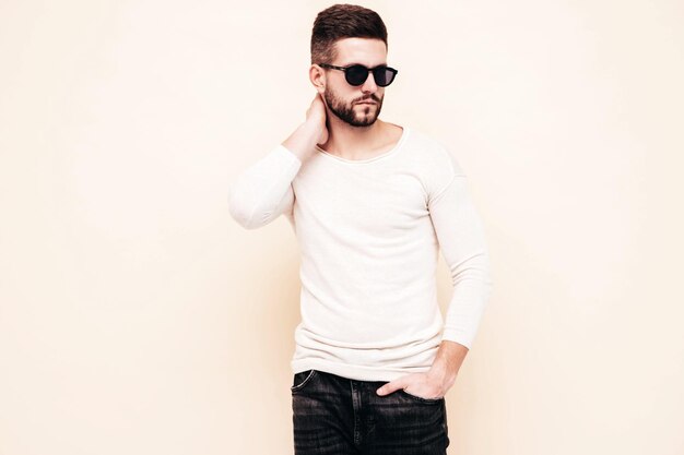 Porträt eines gutaussehenden, selbstbewussten, stilvollen Hipster-Lambersexual-ModelsMann in weißem Pullover und Jeans Modemann, der in der Nähe der Wand im Studio mit Sonnenbrille posiert