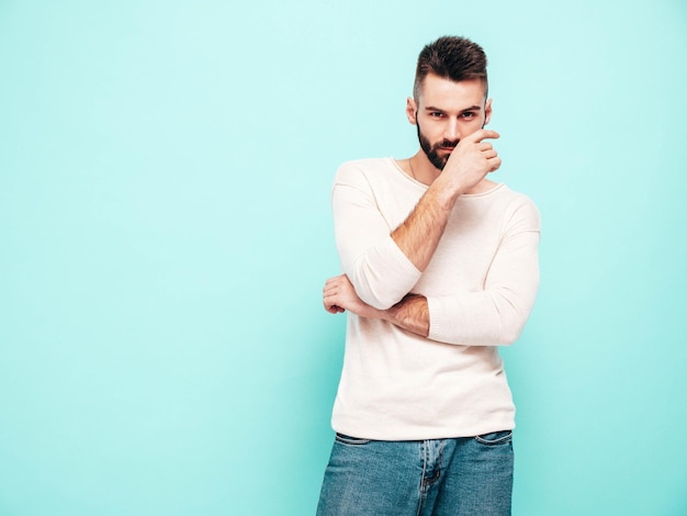 Porträt eines gutaussehenden, selbstbewussten, stilvollen Hipster-Lambersexual-ModelsMann in weißem Pullover und Jeans Modemann isoliert auf blauer Wand im Studio Nachdenklich