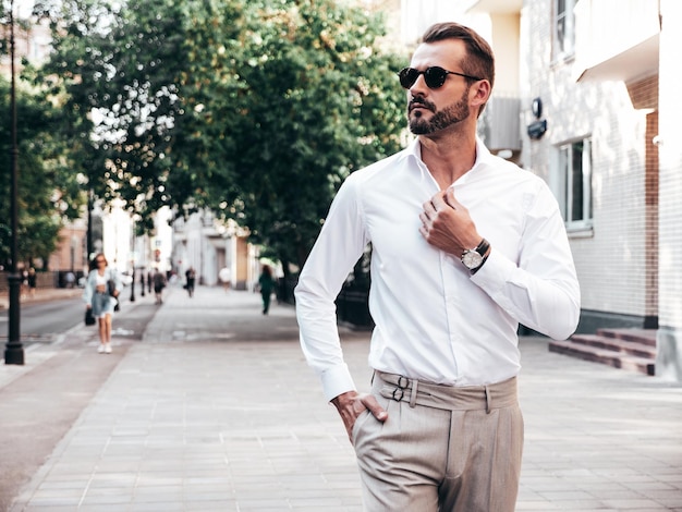 Porträt eines gutaussehenden, selbstbewussten, stilvollen Hipster-Lambersexuellen ModelsSexy moderner Mann in weißem Hemd und Hose gekleidet Modemann posiert auf Straßenhintergrund in Europa-Stadt bei Sonnenuntergang mit Sonnenbrille