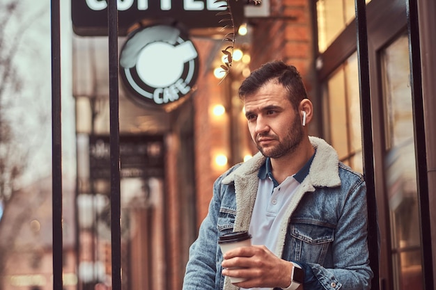 Porträt eines gutaussehenden, stilvollen Mannes, der eine Jeansjacke mit drahtlosen Kopfhörern trägt und Kaffee zum Mitnehmen vor dem Café hält.