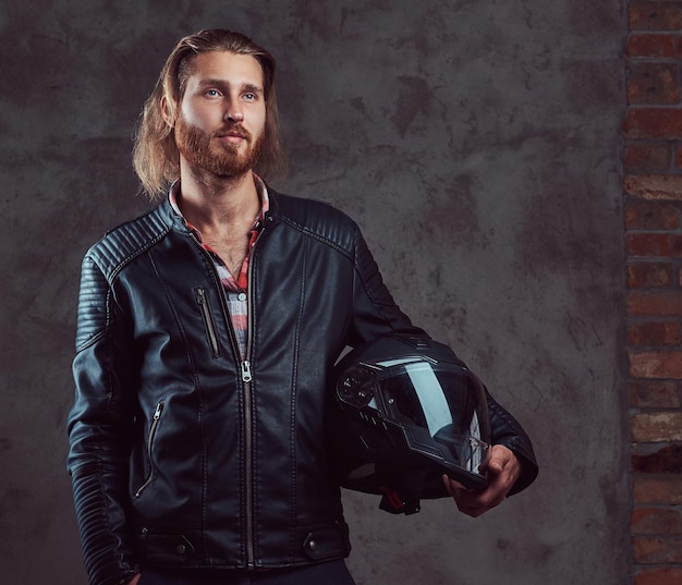 Porträt eines gutaussehenden, stylischen rothaarigen Bikers in einer schwarzen Lederjacke, hält einen Motorradhelm in der Hand und posiert in einem Studio. Getrennt auf einem dunklen Hintergrund.