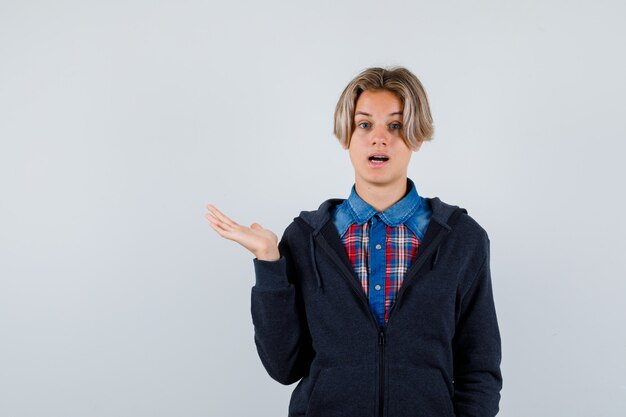 Porträt eines gutaussehenden Teenagers, der die Handfläche in Hemd, Hoodie beiseite ausbreitet und verwirrte Vorderansicht schaut