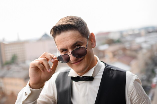 Porträt eines gutaussehenden und glücklichen Mannes, der einen stylischen Anzug mit Fliege trägt, eine Sonnenbrille hält und in die Kamera blinzelt, während er auf unscharfem Hintergrund posiert