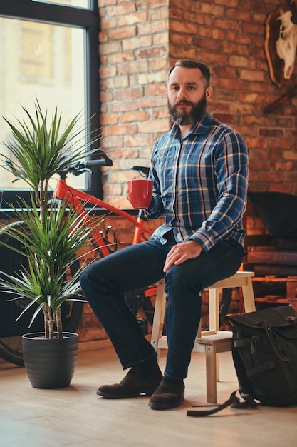 Kostenloses Foto porträt eines hübschen bärtigen hipster-männchens in einem blauen fleece-shirt und jeans hält eine tasse morgenkaffee, während er auf einem holzhocker in einem studio mit loft-interieur sitzt und auf eine kamera blickt.