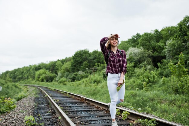 Porträt eines hübschen blonden Mädchens im Tartanhemd, das mit einer Karte in den Händen auf der Eisenbahn spazieren geht