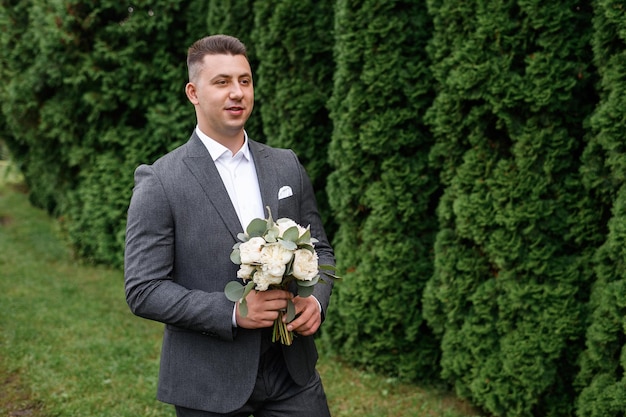 Porträt eines hübschen Bräutigams in einem stilvollen grauen Anzug, der einen Blumenstrauß aus Pfingstrosen hält, der auf die Braut im Garten wartet und geradeaus aussieht Konzept eines attraktiven Verlobten und einer Hochzeitszeremonie