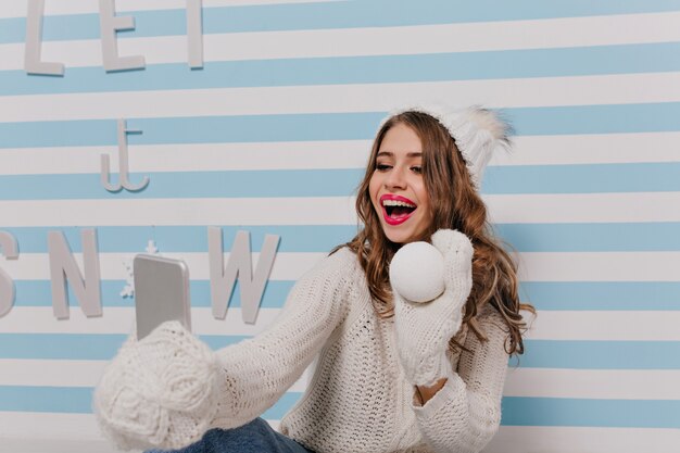 Porträt eines hübschen, gut aussehenden slawischen Modells, das mit Schneeball in ihren Händen aufwirft. Mädchen mit dunkelblonden Haaren in gestrickten Handschuhen macht Selfie