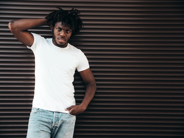 Porträt eines hübschen Hipster-Models. Unrasierter afrikanischer Mann in weißem Sommer-T-Shirt und Jeans. Modemann mit Dreadlocks-Frisur, der in der Nähe der Rollladenwand auf der Straße posiert