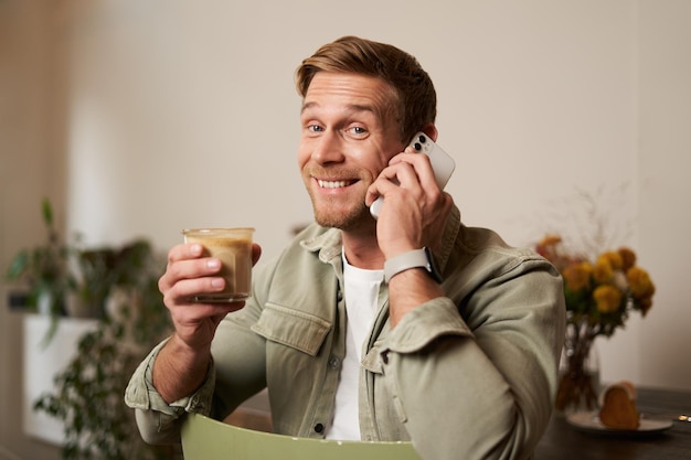 Kostenloses Foto porträt eines hübschen jungen mannes, der am telefon spricht, in einem café sitzt und kaffee trinkt und auf eine anfrage antwortet