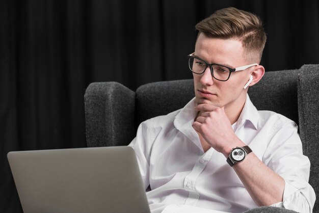 Porträt eines hübschen jungen Mannes, der auf dem Lehnsessel betrachtet Laptop sitzt