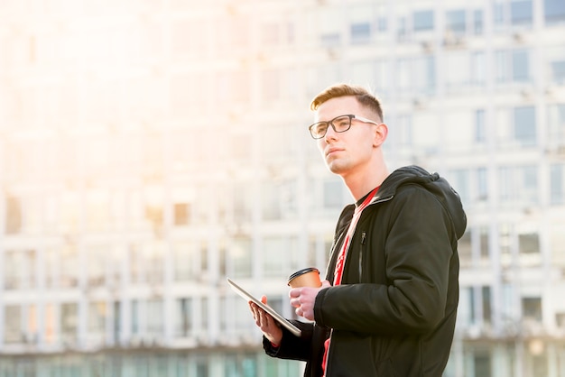 Porträt eines hübschen jungen Mannes, der in der Hand digitale Tabletten- und Mitnehmerkaffeetasse hält