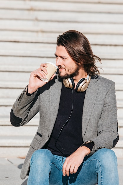 Porträt eines hübschen jungen Mannes mit Kopfhörer um seinen Hals Mitnehmerkaffee trinkend