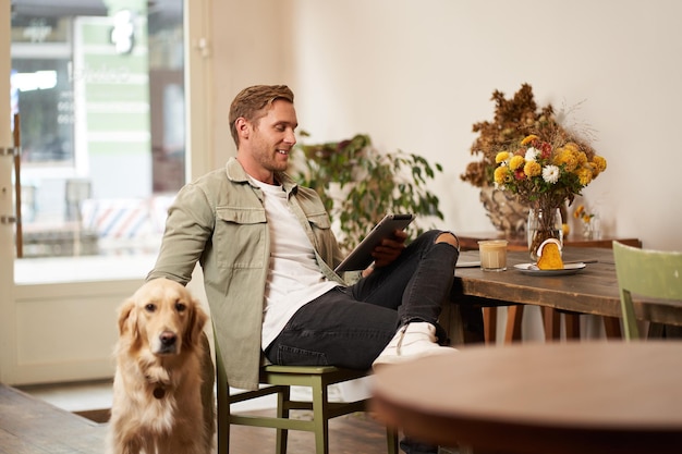 Porträt eines hübschen jungen Mannes sitzt in einem Café mit seinem Hund und streichelt einen Golden Retriever und liest Nachrichten.