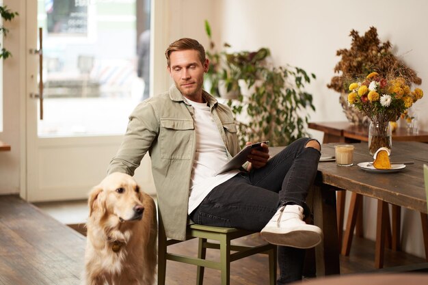 Porträt eines hübschen jungen Mannes sitzt in einem Café mit seinem Hund und streichelt einen Golden Retriever und liest Nachrichten.