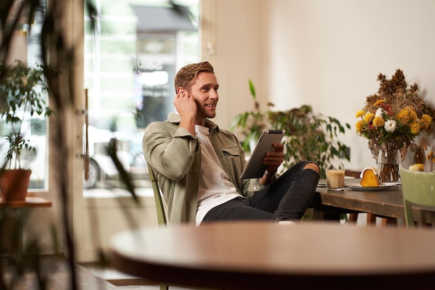 Kostenloses Foto porträt eines hübschen lächelnden jungen mannes, der in einem café sitzt und mit einem digitalen tablet videochats mit