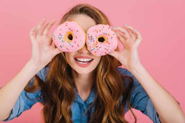 Porträt eines hübschen langhaarigen Mädchens in Retro-Jacke, das mit ihrem Dessert für eine Teeparty spielt, einzeln auf rosafarbenem Hintergrund. Faszinierende junge lockige Frau, die glasierte Donuts als Brille hält und lacht.