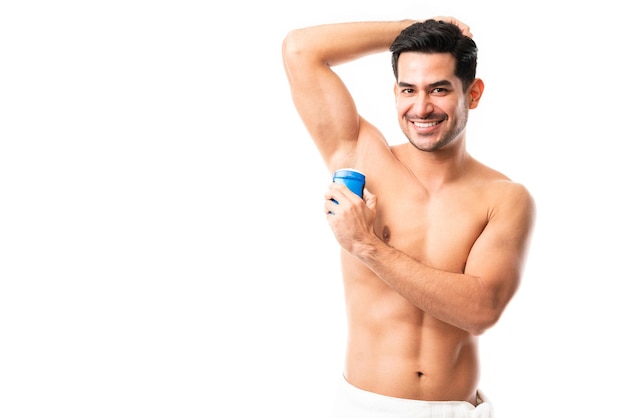 Porträt eines hübschen lateinischen Mannes, der Deodorant hält, während er mit erhobenem Arm auf weißem Hintergrund steht