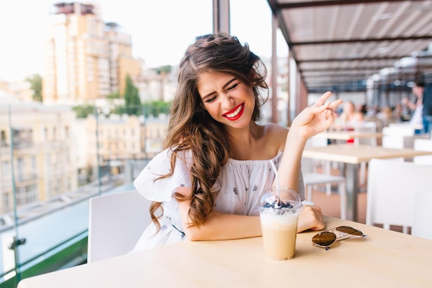 Porträt eines hübschen Mädchens mit langen Haaren, das am Tisch auf der Terrasse im Café sitzt. Sie trägt ein weißes Kleid mit nackten Schultern und rotem Lippenstift. Sie hat Spaß vor der Kamera.