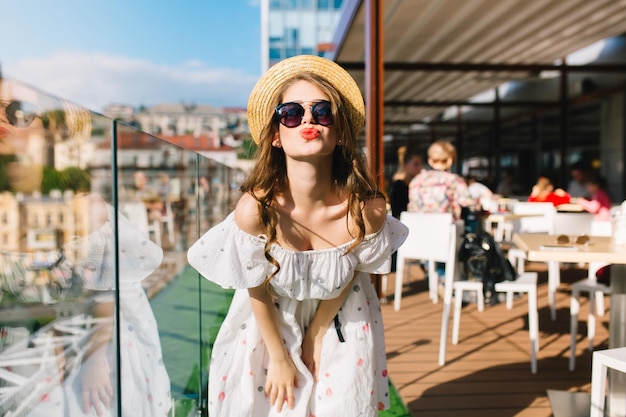 Porträt eines hübschen Mädchens mit langen Haaren in Sonnenbrillen, das auf der Terrasse im Café steht. Sie trägt ein weißes Kleid mit nackten Schultern, roten Lippenstift und Hut. Sie gibt der Kamera einen Kuss.