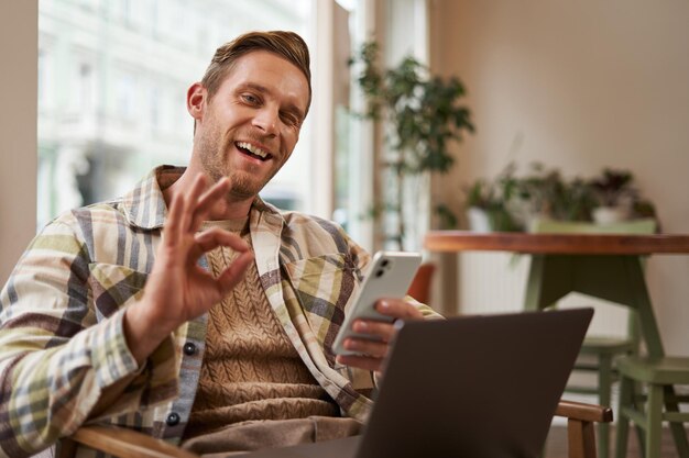 Porträt eines hübschen Mannes in einem Café, der mit einem Laptop sitzt und fern freiberuflich mit einem Telefon in der Hand arbeitet