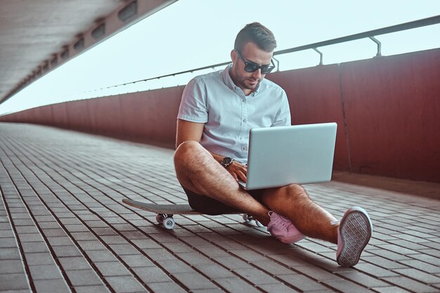 Porträt eines hübschen, modischen Freiberuflers mit Sonnenbrille, gekleidet in ein weißes Hemd und Shorts, der an einem Laptop arbeitet, während er auf einem Skateboard unter einer Brücke sitzt.