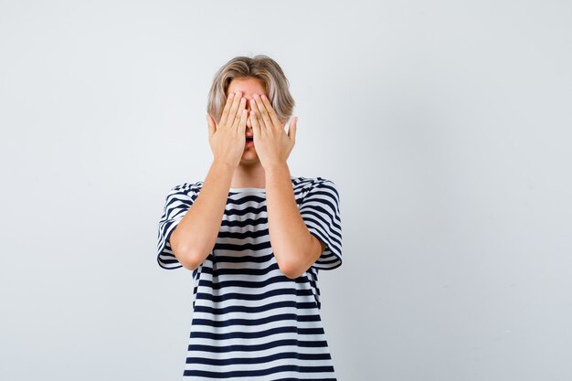 Porträt eines hübschen Teenagers, der das Gesicht mit den Händen im gestreiften T-Shirt bedeckt und verängstigte Vorderansicht schaut