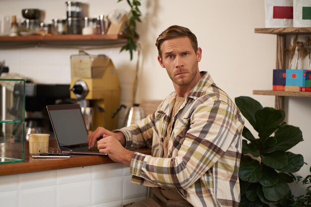 Porträt eines in einem Café arbeitenden Mannes, der verwirrt in die Kamera schaut, sitzt mit Laptop und Kaffeeglas