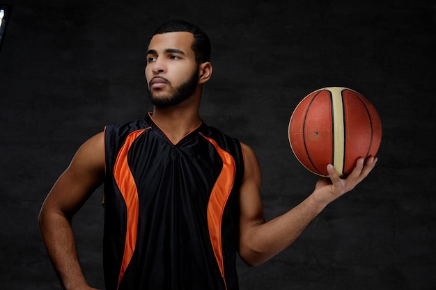 Porträt eines jungen afroamerikanischen Basketballspielers in Sportbekleidung isoliert vor dunklem Hintergrund.