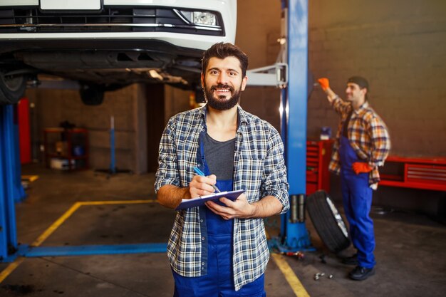 Porträt eines jungen Automechanikers, der in die Zwischenablage schreibt und in die Kamera schaut, während ein anderer Spezialist dahinter arbeitet
